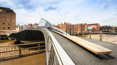 Hull City Council View from Scale Lane Bridge with metal railings, wooden seating and a black tarmac surface  looking towards the brick warehouses along the River Hull 