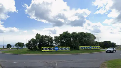 The Highfields Roundabout is a grassed roundabout with trees growing in its centre and signs. A black car is travelling around it. The North Sea is visible in the distance.