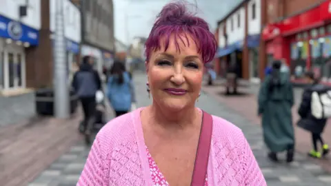 A woman with purple hair, pink cardigan and pink handbag