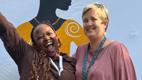 Grit Two people smiling and posing together in front of a colourful background featuring artwork of a silhouetted woman.