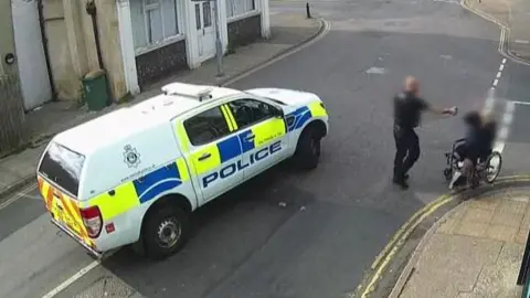 A police truck parked in the middle of King Street, Great Yarmouth, with the police officer, arm extended, deploying PAVA spray on a man, who is in his wheelchair on the white line of the junction of St Peter's Road. Behind the police van is the Old White Lion pub, which is converted into flats, and to the right of the image is the frontage of the Londis convenience store. In the background is King Street looking towards Great Yarmouth town centre.