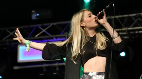 A woman with blonde hair is singing into a microphone which she holds with her left hand, her right hand is outstretched. She wears a black jacket over a black crop top. She is on stage. The woman is Violet Skies.