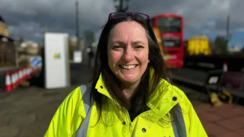 Pamela Holmes from Newcastle City Council wearing a high viz jacket standing in front of the Tyne Bridge with a double decker bus going past 