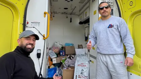 Supplied Jake Trask and Rhys Edwards smiling while looking into the camera. They are standing in front of the back entrance to an ambulance, which is filled with supplies, dog food, brown boxes and other items.
