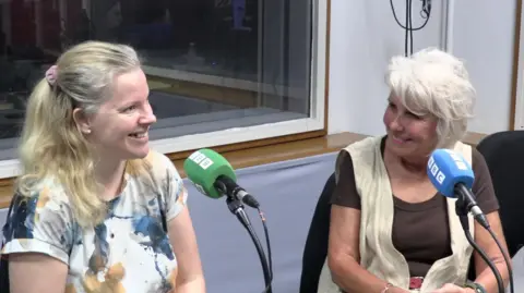 Ellie Mead and Helen Russell sit in front of microphones in a BBC radio studio. Ellie has long blonde hair in a pony tail and a mainly white top. Helen has shorter grey hair and wears a light waistcoat over a brown top.