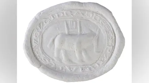 Andrew Williams/Norfolk County Council An impression in white wax of an oval seal matrix. It shows a narrow band on its circumference which is inscribed with letters. Its middle shows a portly-looking elephant with broad legs and tail and a trunk. On its back is a castle or howdah.