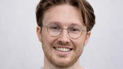 Fergal Mackie smiling at the camera. He has brown hair and is wearing glasses.