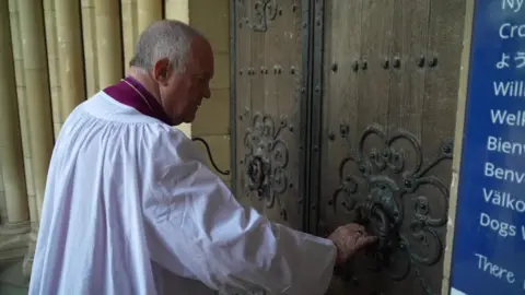 BBC Bishop Williams, wearing white purple clerical robes, takes hold of a large door knocker on the front door of one of the large, ornate entrances into Truro Cathedral.
