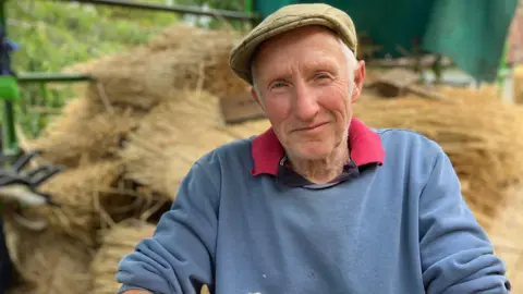 An older man is smiling at the camera. He is wearing a tweed cap, a pale blue jumper with a red polo top collar poking out the top. Behind him are piles of straw. 
