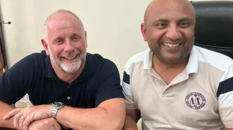 Two men smile at the camera. One has a a short white beard and is wearing a blue polo shirt. The other has no hair or beard and is wearing a white polo shirt