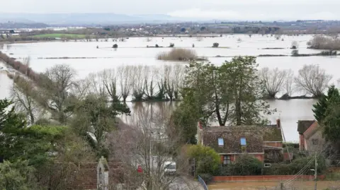 A number of flooded fields are seen from above. Two houses are visible in the foreground, surrounded by water. 