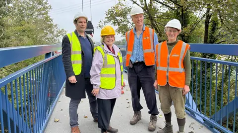 Councillors Richard Wilkins and Ros Wyke wearing yellow hi-vis vests and hard hats, standing with Strawberry Line volunteers on the new footbridge over the B3136 West Shepton, in Shepton Mallet. 