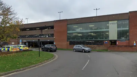 A large multi storey car park has four floors and has cars parked outside, including two police cars which are on the left. There is a small area of grass on the left and in front of the building.