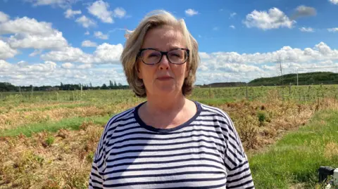 Emma Howgego/BBC A woman wears a white and black stripped top. She has chin length blonde hair and glasses. In the background are some overgrown allotment plots. 