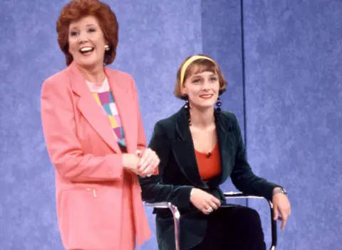 Shutterstock/ ITV Frankie (R) is smiling and sitting on a chair next to Cilla Black, who is standing. Frankie is wearing a bottle green blazer over an orange-red top. Her blonde hair is styled in a short bob with a fringe and held back by a bright yellow hairband. Cilla is wearing a peach jacket over a checked, pastel top, with red nail varnish. Her signature auburn hair is worn in a shoulder-length bouffant style.