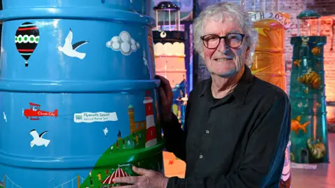 St Luke's Hospice Plymouth/Paul Slater Images Brian has grey curly hair and large glasses. He is standing next to a blue lighthouse featuring Plymouth scenes