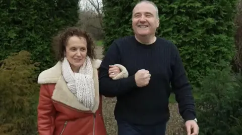 Lee Castleton, wearing a knitted black jumper, walks arm in arm with wife Lisa, who is wearing a red leather coat with wide fleece collar. They are walking along a gravel path, with tall conifer trees behind them.