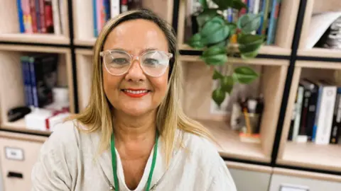 Emma Cunningham Dr Emma Cunningham. She has shoulder-length blond hair and is wearing glasses with a large, round, clear rim. She is wearing a beige top with a green lanyard around her neck. She is wearing red lipstick and smiling. Behind her, and slightly out of focus, are bookshelves made from cream wood cubes. On one there is a plant.