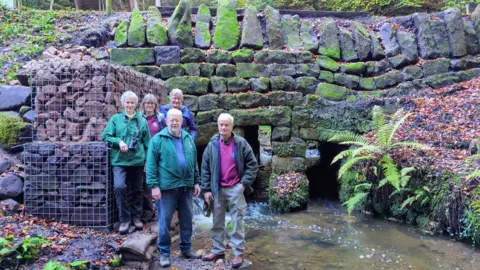 Five people stand next to a stream. Behind them is a stone bridge covered in moss. Two large cages containing large rocks are visible on the left behind the group.