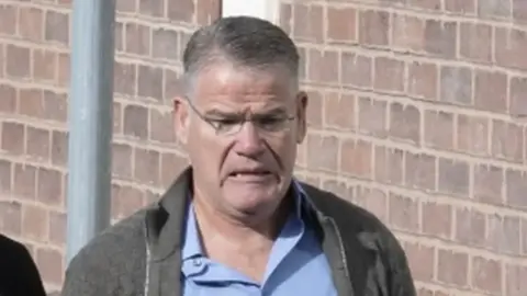 A man with grey hair, glasses and a grey cardigan standing near a brick wall.