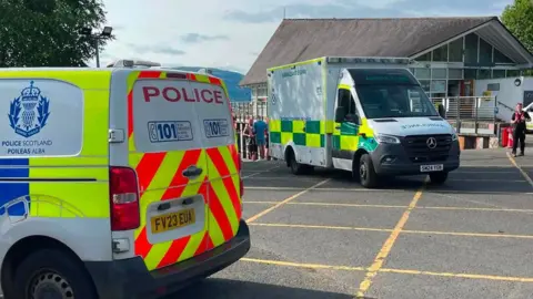 Loch Lomond Rescue Boat A police van and an ambulance in a car park outside a building with railings. A hill is visible in the background.