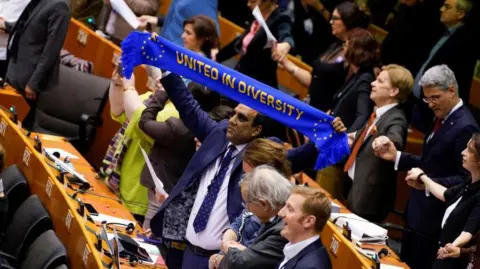 Getty Images Baron Shaffaq Mohammed stands in the European Parliament during his time as an MEP. He is holding a blue scarf that says 'United in Diversity'. It also features the EU flag. 