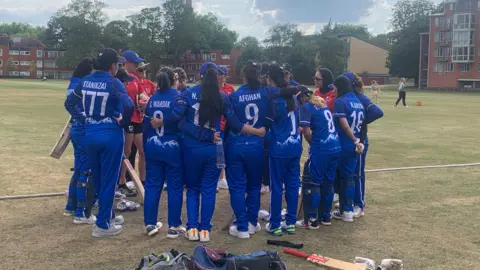 Louise Hulland The women's team group together in a huddle, wearing their royal blue kit with white writing. They stand next to the cricket pitch with some holding bats. There are residential buildings and trees in the distance.