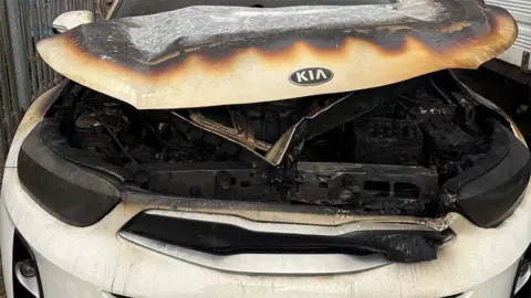 Sian Crooks A close up of the fire-damaged engine. It is completely black and partly covered by the bonnet of the white car which is bent and burned