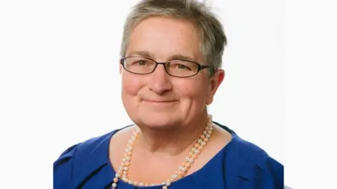 Head and shoulders shot of Councillor Cheryl Payne. She is wearing a blue top, a necklace and spectacles. She has short hair.