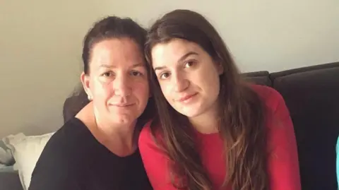 Una Srabovic-Ryan Una is sitting on a sofa next to her mother. Una is wearing a red top, while her mother is wearing a black top.