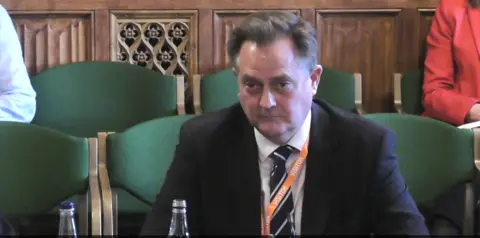 parliamentlive.tv A man with a suit and tie and short brown hair sits in front of a committee 