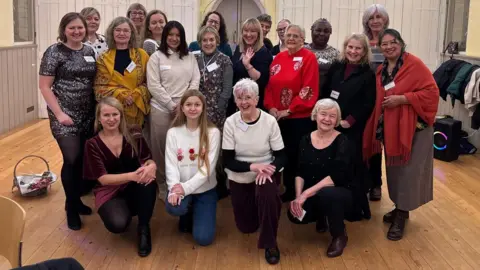 Cad Taylor/BBC A large group of women stand in a room smiling. Some are kneeling at the front, other standing at the back