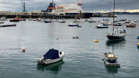 Looking over St Peter Port harbour there are small motor and sail boats and a Condor Ferry moored to the harbour wall in the distance.