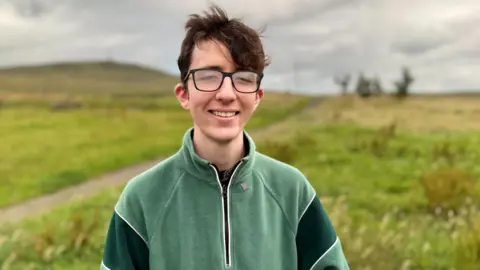A young man with short brown hair, wearing glasses and a green fleece. He is standing in a field with a grey sky above.