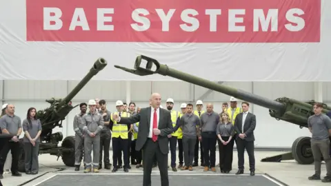A man in a smart suit and red tie stands delivering a speech in front of a large group of people in hard hats and safety jackets. They all stand under two large artillery pieces.