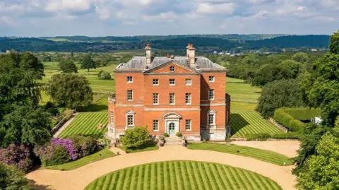 Mo Chaudry An aerial shot of a large hall with a circular area of lawn in front of it and two rectangular areas of lawn visible either side of it. There are steps leading to a green door which has to tall windows either side. There are further tall windows on the second floor, with smaller windows on the third floor and a small circular window at the top of the building. There is rolling countryside in the background behind the building.