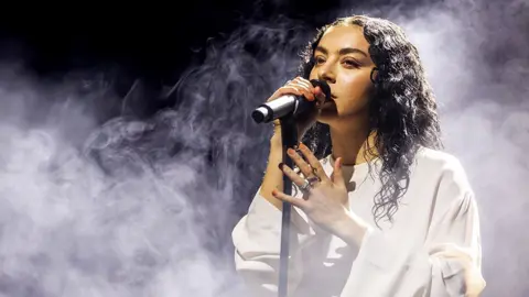 Getty Images Charli XCX, a female singer performing on stage, singing into a black microphone while wearing a white outfit, while smoke effects surround her.