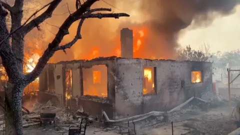 The shell of a home with flames around it 