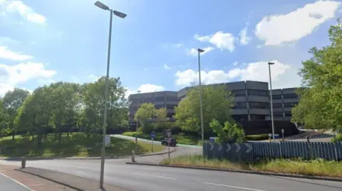 Google A large grey building, with three four-storey towers visible rises behind large green trees and a grassy area to the left. A two-lane main road runs from left to right in front of the site