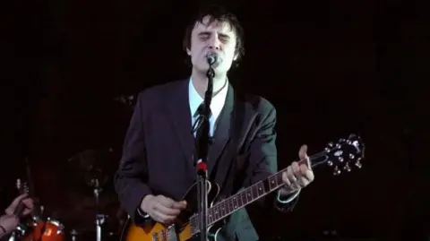 Getty Images Pete Doherty stands on stage singing into a microphone and playing an electric guitar while performing with The Libertines during The Libertines in Concert at Brixton Academy in London in 2004. He has brown hair and is wearing a black suit with white shirt and black tie. The corner of a drum kit can be seen behind him to the left. The background is black. 