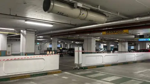 Scene of incident at Charlestown Shopping Centre underground car park in Finglas. There is red and white tape in the foreground as well as white walls. There is a yellow and black sign saying caution near the roof.