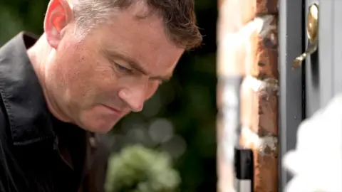Ed Thomas leans in at a front door, to listen to the opinions of Mary through her doorbell speaker.
