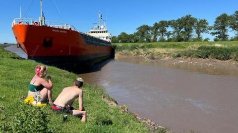 Couple on river bank look at stranded ship