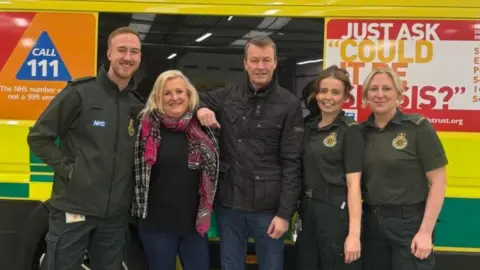 A man and a woman are standing alongside three ambulance crew members in uniform. They are standing by the side of an ambulance with West Midlands Ambulance Service livery.