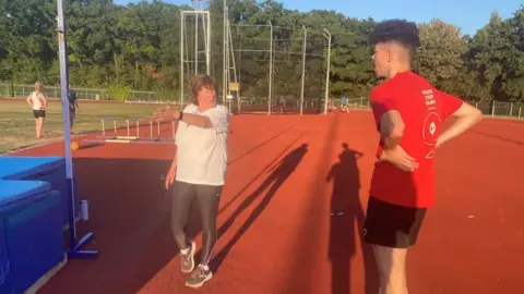England Athletics A woman in a white t-shirt and black sports leggings coaches a young athlete wearing a red top and black sports at an athletics track.