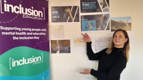 Inclusion Education Marie Greenhalgh pointing to a sign that says: Inclusion Education: supporting you people with mental health and education the Inclusion Way. There are also pieces of paper promotion the Baton of Hope Tour 2025 behind her on a wall