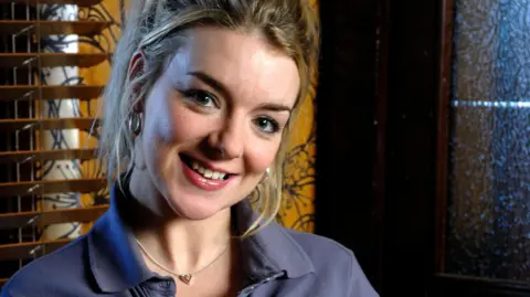 BBC Sheridan Smith. She has blonde hair and smiles as she looks into the camera. She is standing in front of a wooden blind and a wall covered in amber and black wallpaper. She is wearing a blue polo shirt, silver hoop earrings and a silver pendant necklace.
