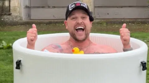 Image of Harry Beattie in a round ice bath, smiling at the camera with his thumbs up, all he is wearing is a black cap.
