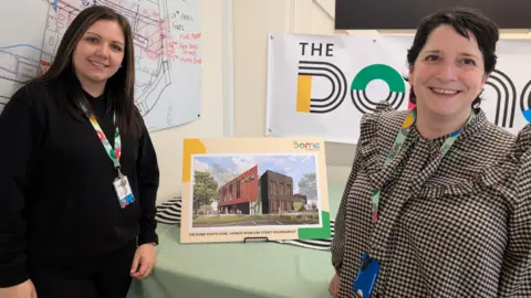 Kelly Prince on the left and Ellen Birchenough on the right, with a CGI image of the completed building on a board in between them both. Prince has long dark hair and wears a black top and trousers - Birchenough has a black and white checked top and short dark hair.