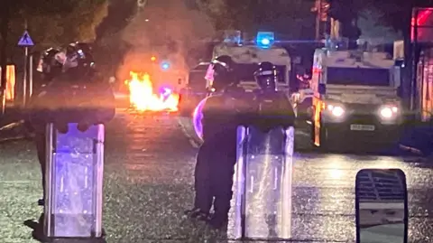 BBC Police standing with riot shields in front of a fire during disorder